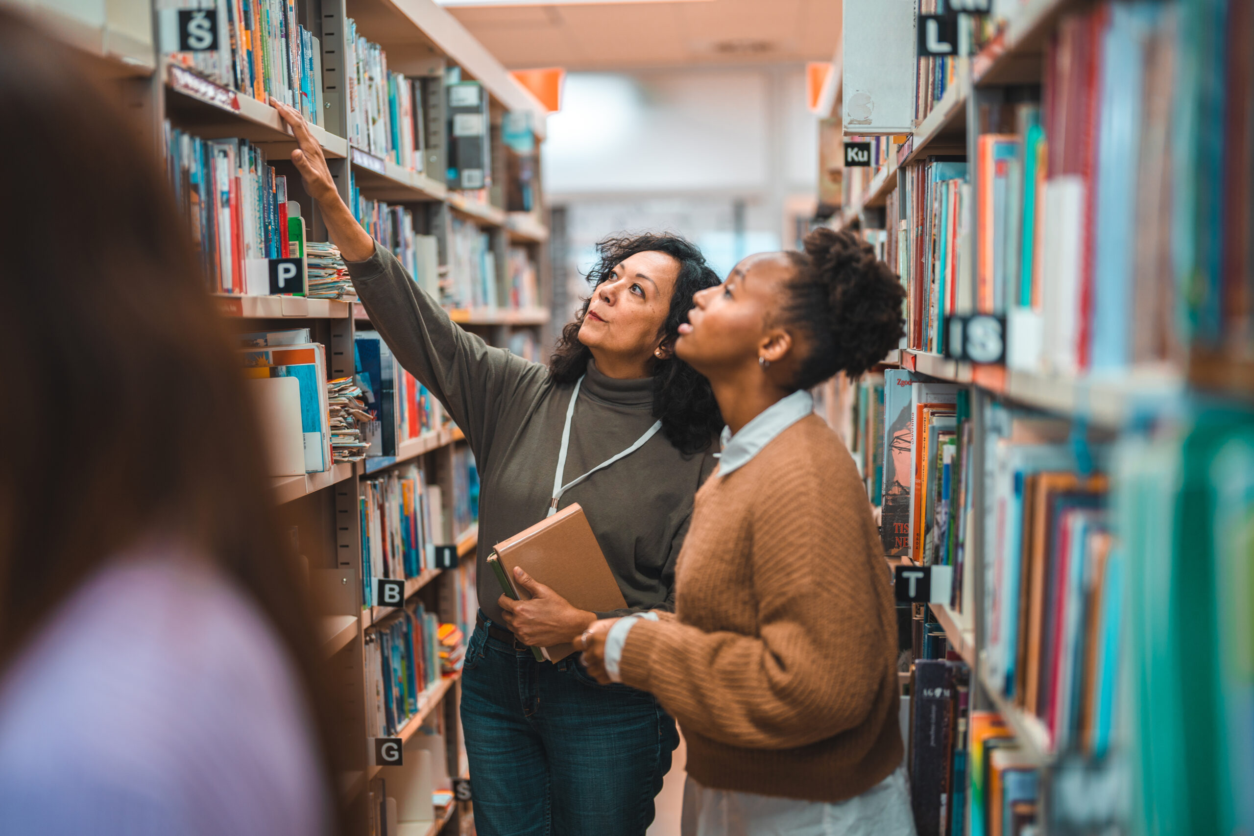 Student searching for a specific book in a library. Walking in between the book shelves and smiling.